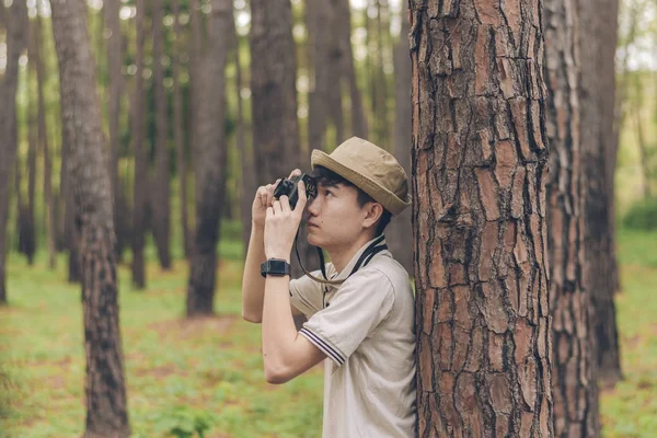 Asia man wears shirt, and hat are stand against the tree and taking photos at the forest.