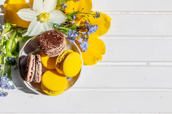 macarons veya Bademli kurabiye tatlı tatlı yemek güzel. Beyaz ahşap bir masa üzerinde çikolata ve limon tatlılar. Lezzetli tatlı düz lay ve nergis ve unutma çiçekler.