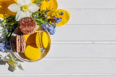 Beyaz ahşap masa üzerinde plaka üzerinde renkli kurabiye. Tatlı macarons ve çiçekler. İhale ile kompozisyon çiçekleri çiçek ve tatlı Fransız. Kopya alanı ile üstten görünüm