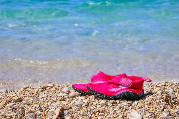 Pair of pink swimming shoes on marble pebble beach beside turquoise sea water. Summer holiday concept. Kids shoes for beach.Swimming neoprene shoes.Water shoes, rocky beach — Stock Photo, Image