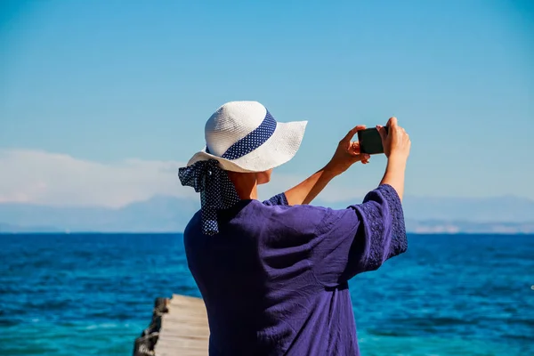 Hermosa chica en sombrero de verano blanco y camisa azul en la playa hace selfie, foto. Historias de vacaciones por internet. Vacaciones de verano en el mar. Clima soleado. Copiar espacio — Foto de Stock