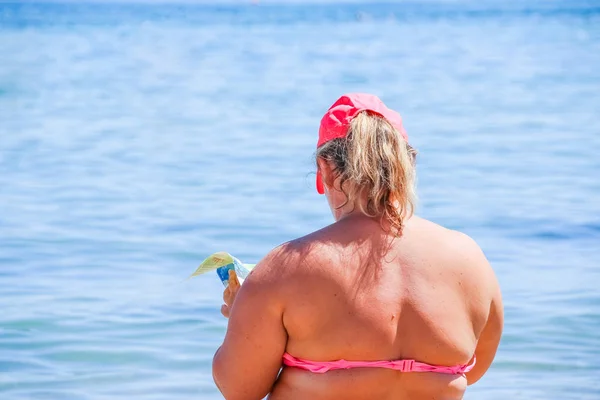 Mature woman in swimwear at the sea. Overweight woman in swimsuit comes on the beach, reading a book, rear view.Fat women sitting on a beach by an ocean — Stock Photo, Image