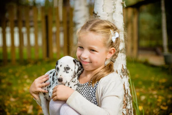 Little cute blonde girl playing with her Dalmatian puppy outdoo, on sunny warm autumn day.care of Pets concept. Child kisses,hugging her dog, the concept of childrens and dog emotions, of friendship