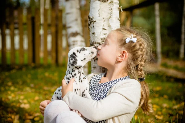 Söt tjej kysser hennes valp, doggy på trä staket bakgrunden. Glad tjej med en hund slickar hennes face.real vänner. Söt dalmatiska valp kyssar söta blonda hår flicka. positivt tänkande och känslor — Stockfoto