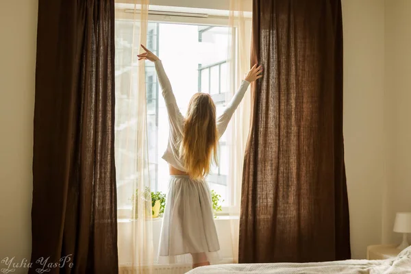 La mano abre la cortina en la ventana de la casa de madera. La brillante luz del sol brilla a través. Hermosa luz de la mañana soleada detrás de la ventana del chalet. espalda de mujer silueta — Foto de Stock