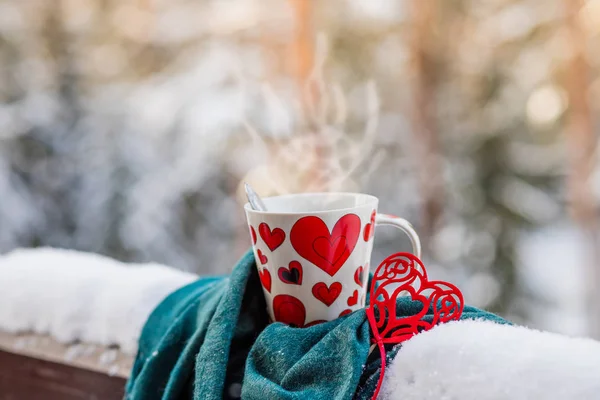 Белая чашка кофе на заснеженном балконе, патио. Зимним утром на курорте Кубок с красными сердцами и горячим напитком, завернутым в шарф в снежный символ любви. Концепция — стоковое фото