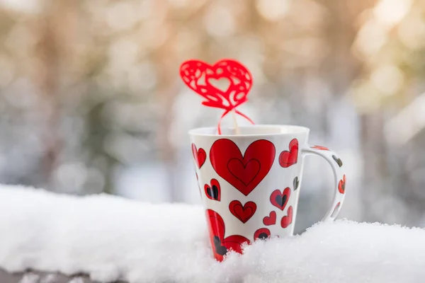 Love at coffe time, christmas concept, mug and red heart decoration on a snow balcony.Cup with heart and a hot drink wrapped in a scarf in the snow.Valentines day concept — Stock Photo, Image