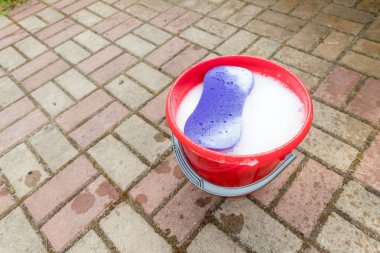 Mor renkli sünger ile arnavut kaldırımlı arka plan üzerinde kırmızı plastik kova. Temizleme teması. Ev içi arka planda ev aletleri, yay temizliği, toparlama, temizlik hizmeti konsepti
