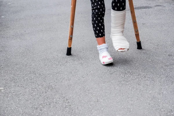 Young girl in orthopedic cast on crutches walking on the street near the road. Child with a broken leg on crutches, ankle injury. Bone fracture and ankle fracture in children