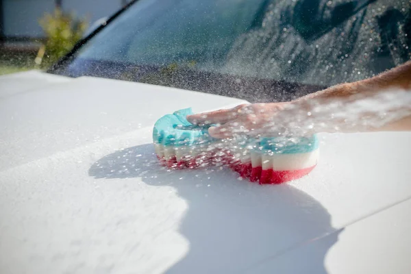 Człowiek mycie mydłem biały samochód z kolorową gąbką. Ręczny Myjnia samochodowa w myjni samochodowej z pracownikiem pracownika. mycie samochodu w słoneczny dzień — Zdjęcie stockowe