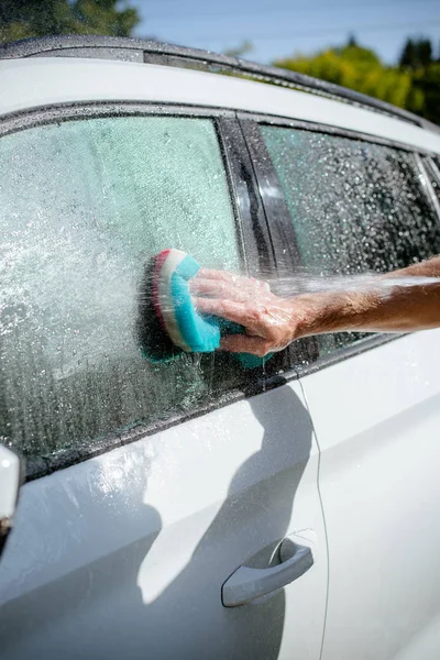 Man tvättar en tvål vit bil med en färgglad svamp. Manuell biltvätt i biltvätt Shop service med anställd arbetstagare. bil tvättanläggning på solig dag — Stockfoto
