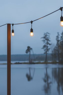 Alacakaranlık, lakescape gri renkli gökyüzü, çelenk ve ahşap teras, iskele olarak elektrikli lambalar. akşam romantik bir atmosfer. Gece kafe terası. Ev dekorasyonu için Retro çelenk.