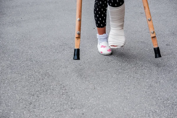 kid with broken leg is on crutches on the street. conceptual photo depicting a child with a broken leg on a holiday, on the school holiday.Girl injured in feet has bandage with crutches on asphalt