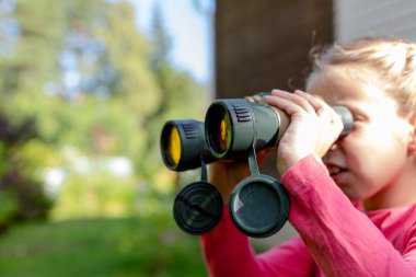 Sarışın küçük kız dürbünle doğa geçmişini inceliyor. Bebek yeşillikte saklandı ve pusuda bekliyor. Çocuk Macera Konsepti. Çocuk uzaktan ebeveynleri izliyor.