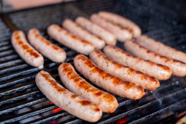 Sosisler ızgarada, dumanlı. Pork sosisleri portatif bir barbeküde ızgara yapıyor. Bir sosis yaz pikniğinde bir çift maşaya konuyor.