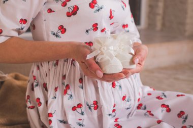 Kadının elinde doğmamış bebek için küçük ayakkabılar. Bebek için bir çift beyaz ayakkabı. Aqua menthe elbiseli hamile kadın karnını ve bebek ayakkabılarını tutuyor. Hamilelik, annelik ve beklenti konsepti.