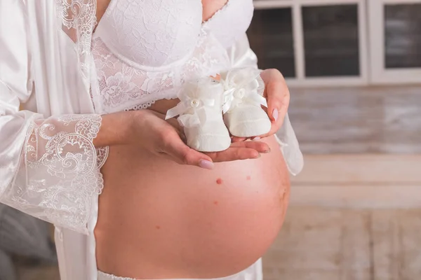 Sapatos pequenos para o feto na mão da mulher. Par de sapatos brancos para bebê. Mulher grávida em aqua menthe vestido segurando sua barriga e os sapatos de bebê. Conceito de gravidez, maternidade e expectativa. — Fotografia de Stock