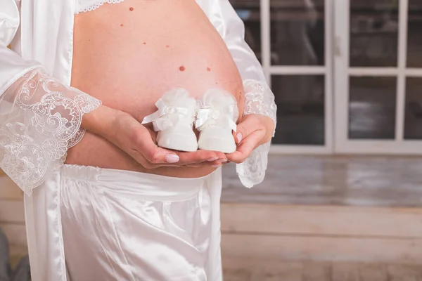 Sapatos pequenos para o feto na mão da mulher. Par de sapatos brancos para bebê. Mulher grávida em aqua menthe vestido segurando sua barriga e os sapatos de bebê. Conceito de gravidez, maternidade e expectativa. — Fotografia de Stock