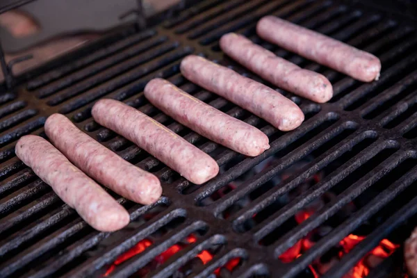 Surowe kiełbaski wieprzowe grillujące na grillu na letnim pikniku, zamykanie grilla, mięsa i ognia.Grillowane kiełbaski na grillu z dymem i płomieniem na łące. Piknik grillowy.Skupienie na fakultatywnym — Zdjęcie stockowe