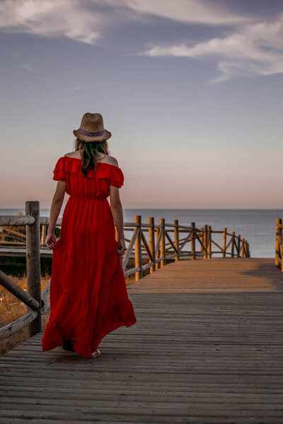 A female model with her back turned walking down a wooden path to the sea during sunset