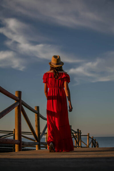 A female model walking away down a path towards the sea in a red dress
