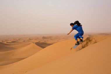 Fas'ta Erg Cheggaga kumulları üzerinde kum tahtası sürme sırasında Touareg Gandoura performans atlama hile sandboarder giymiş yan görünümü 