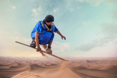 sandboarder dressed in Touareg Gandoura jumping while riding sand board on dunes of Erg Cheggaga in Morocco  clipart