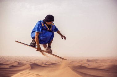 sandboarder dressed in Touareg Gandoura jumping while riding sand board on dunes of Erg Cheggaga in Morocco  clipart