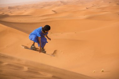 sandboarder dressed in Touareg Gandoura jumping while riding sand board on dunes of Erg Cheggaga in Morocco  clipart