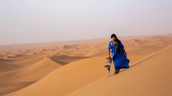 Touareg Gandoura'da kum tahtası tutan ve Fas'ta Erg Cheggaga kumulları arasında yürüyen sandboarder görünümü 