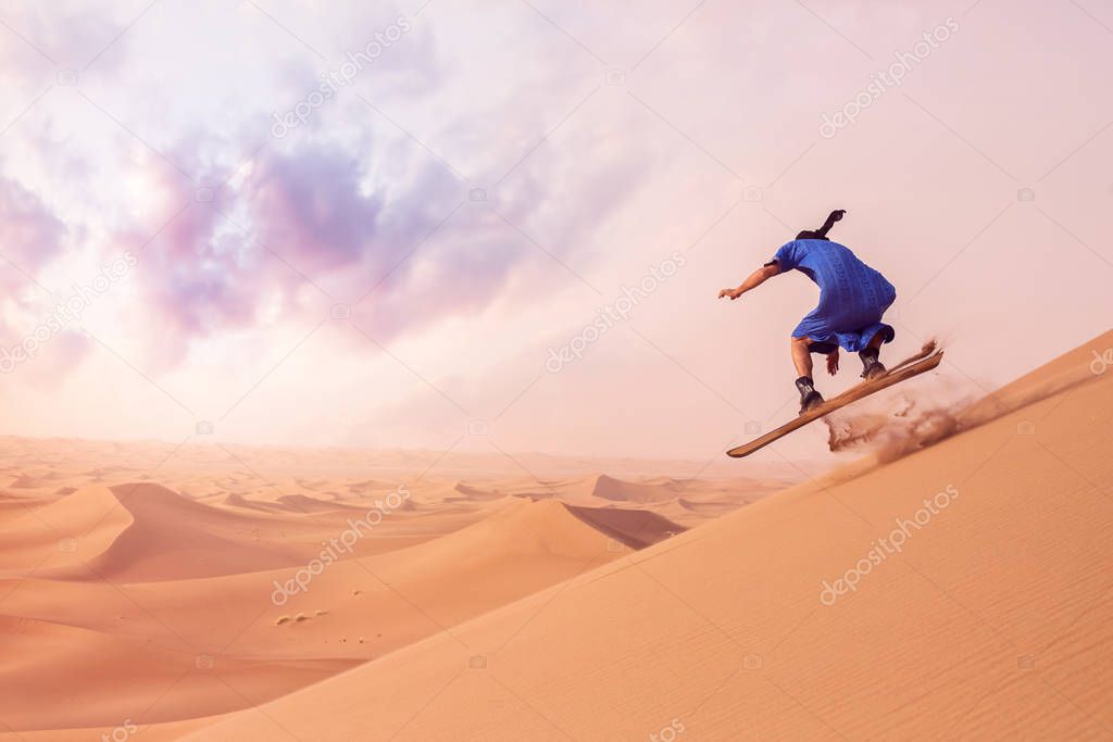 back view of sandboarder dressed in Touareg Gandoura performing jump trick while riding sand board on dunes of Erg Cheggaga in Morocco 