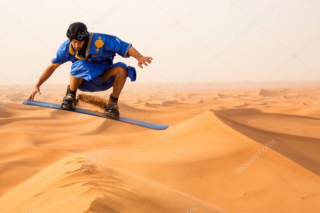 sandboarder dressed in Touareg Gandoura jumping while riding sand board on dunes of Erg Cheggaga in Morocco 