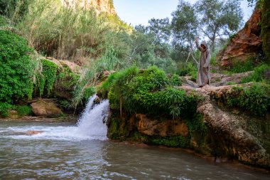 ormanda kayalıklar arasında çağlayan ile dağ nehri kıyısında duran baston ile Arap tarzı bornoz adam, Fas 
