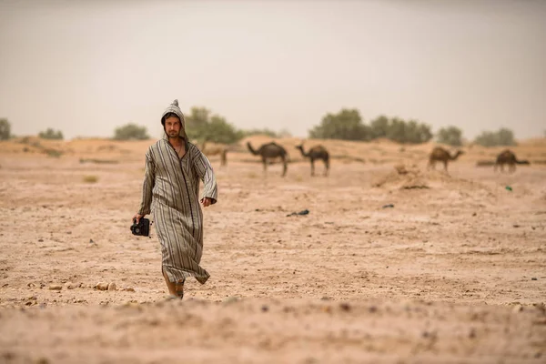 man in  in arabic style robe walking in sandy desert with camels on background, Morocco