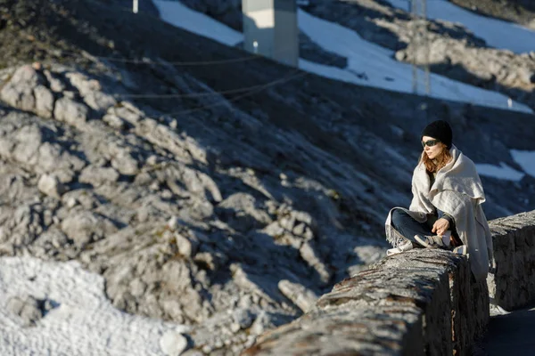 Dívka sedí na pozadí hor. Na pozadí zasněžených hor v klobouku a slunečních brýlích. — Stock fotografie