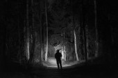 Mann, der nachts im Freien in einer Baumallee steht und mit Taschenlampe leuchtet. schöne dunkle, schneebedeckte Winternacht.