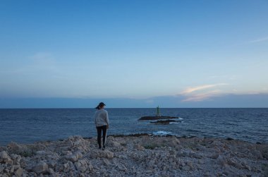 Genç bir kadın gün batımında sahilde duruyor.