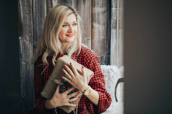 Glückliche junge blonde Frau im roten Pullover erhielt ein Geschenk zu Weihnachten, Neujahr, das Mädchen lächelt und hält eine Geschenkbox an ihrer Hand — Stockfoto
