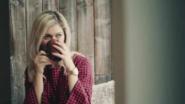 Hermosa chica rubia que mira a la ventana con una taza de café en las manos — Vídeos de Stock