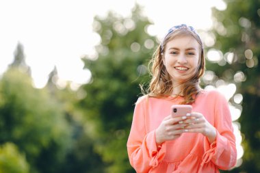 Parkta akıllı telefonlara bakan güzel bir genç kız. Arka planda yeşil ağaçlar olan kameraya bakan güzel bir kadın. İletişim ve doğa kavramı. Ön görünüm.