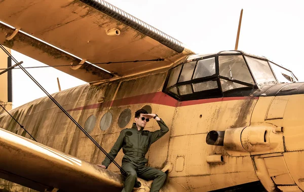 A handsome young pilot sitting on the wing of a plane and coveri — Stock Photo, Image