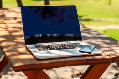 Güneşli bir günde ev ofisinin ya da ev okulunun bahçesindeki bir masada dizüstü bilgisayarlı bilgisayar, akıllı telefon ve kahve fincanı görüntüle.