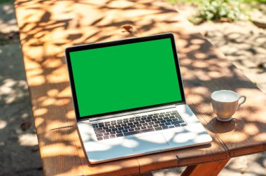 Güneşli bir günde ev ofisinin ya da ev okulunun bahçesindeki bir masada yeşil ekranlı ve kahve fincanlı bir bilgisayar ekranında görüntüle.