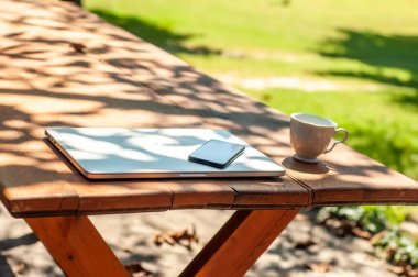 Kapalı bir bilgisayarda ve güneşli bir günde evde ya da evde okul ortamında bahçedeki masada duran bir akıllı telefon kahve fincanına bakın..