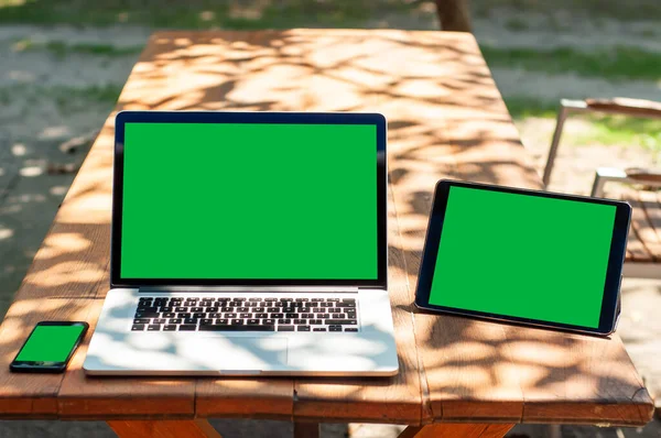 Bir dizüstü bilgisayar, akıllı telefon ve yeşil ekranlı tablet bilgisayarla güneşli bir günde evde ya da evde okul ortamında bahçedeki bir masada görüntüle.
