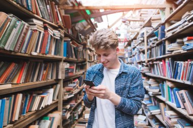 pozitif genç adam bir smartphone bir halk kütüphanesi ve gülümsüyor kullanır. Ellerini rahat kitaplığında bulunan bir telefon ile bir öğrenci portresi.
