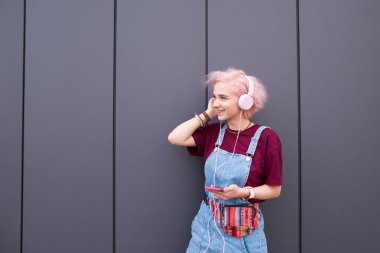 Renkli saç ve denim sarafan ile mutlu, trendy kız müzik pembe kulaklık kulaklık ve gülümsüyor müzik pembe saçlı Genç kız dinler dinler.