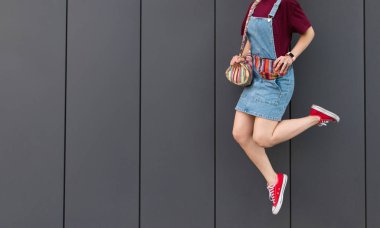 Karanlık bir duvar arka planı atlar şık bir kız kot sarafan ve kırmızı spor ayakkabılar içinde başsız portresi. Karanlık bir arka plan üzerinde atlama bir kızın moda portre.