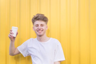 Bir fincan kahve arka planda sarı bir duvar ile ayakta ve gülümseyerek mutlu bir genç adam.