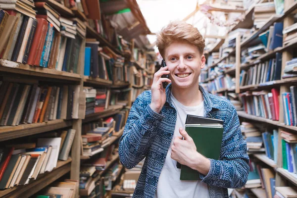 公共図書館で彼の手の本と立っていると電話で話して幸せな若い男の肖像画 笑みを浮かべてその学生は居心地の良い図書館に電話で話す — ストック写真